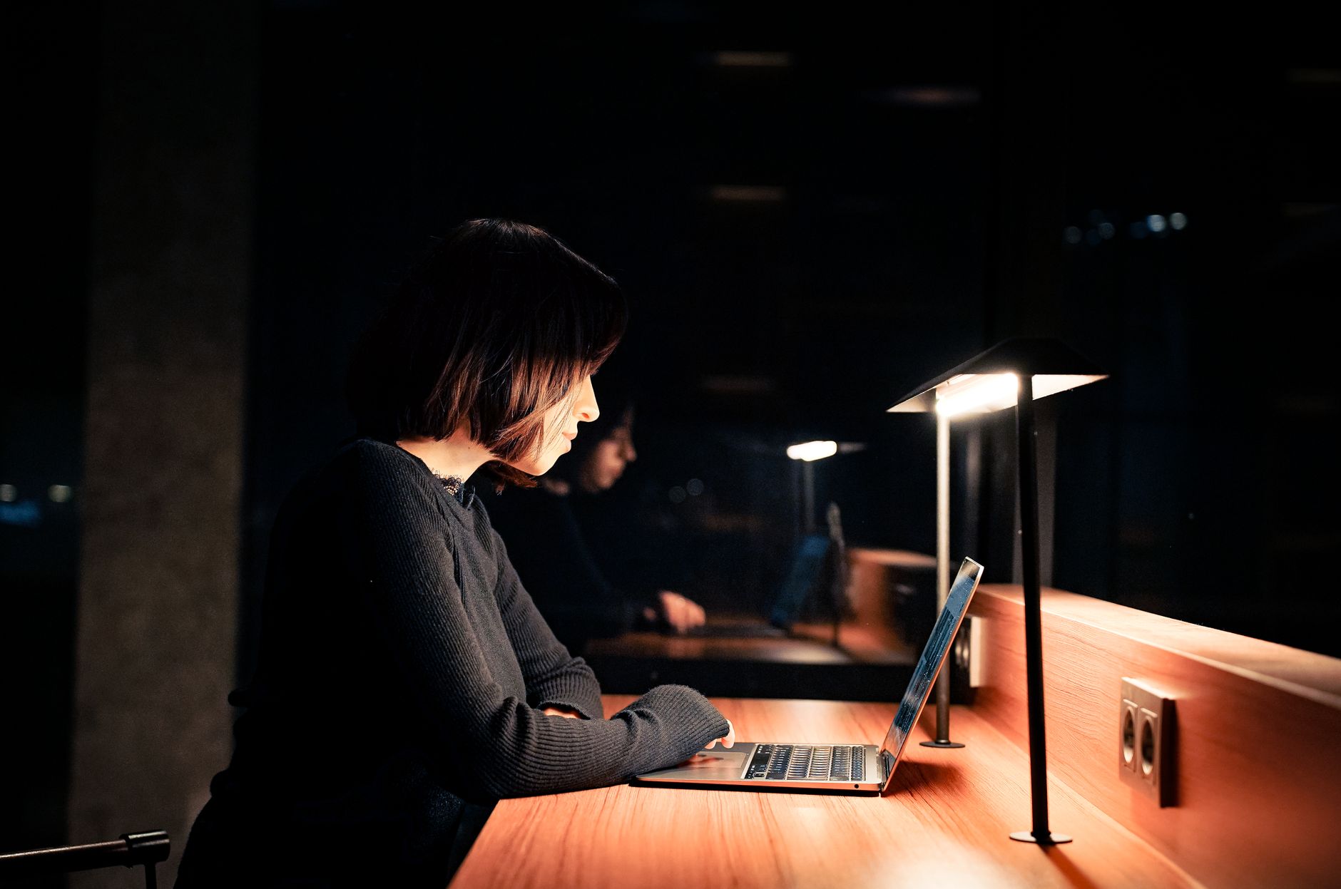 Une femme de profil travaille sur un ordinateur portable à un bureau en bois, éclairée par une petite lampe dans une pièce sombre.