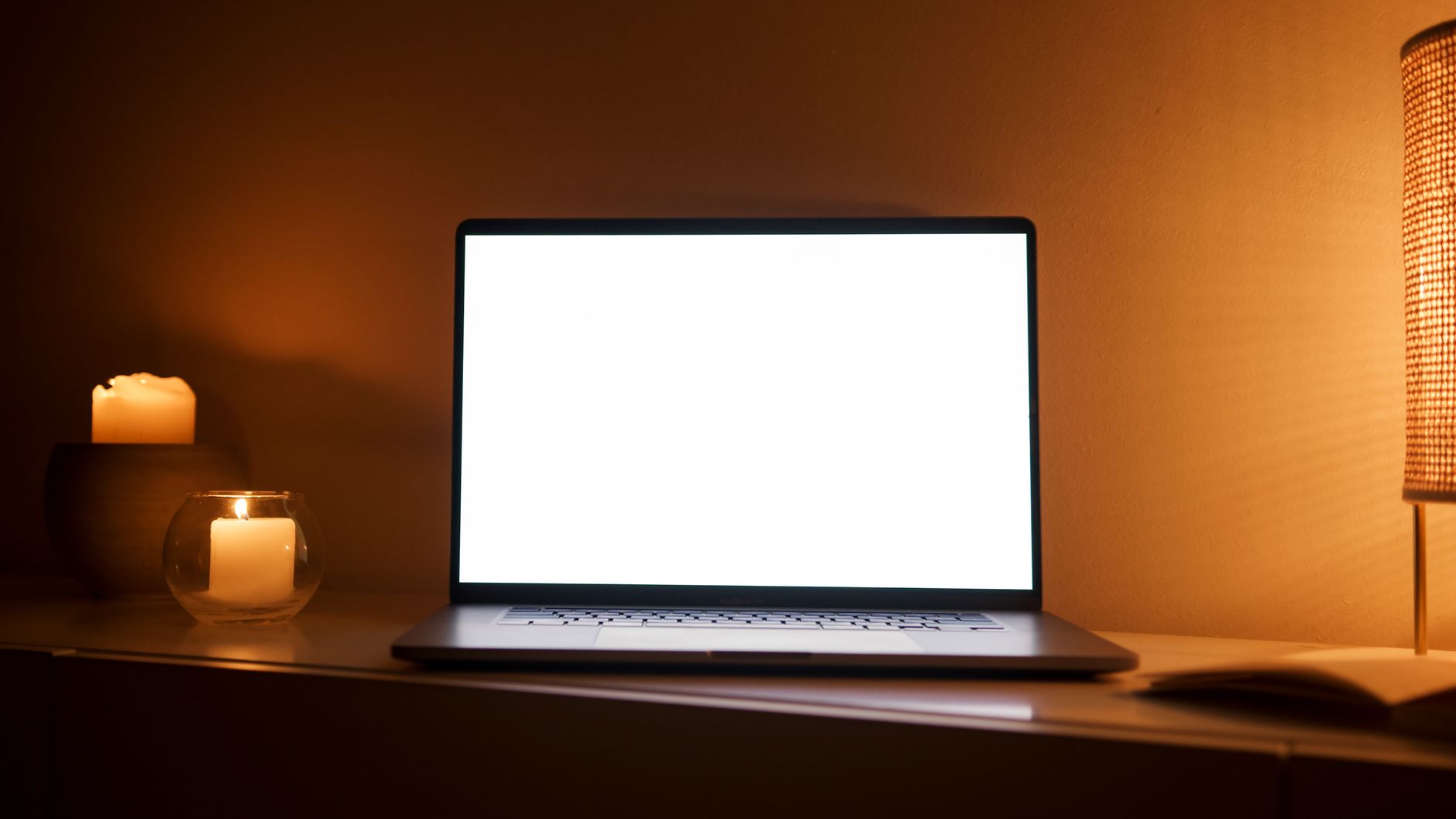 Un ordinateur portable ouvert sur un meuble, entre des bougies allumées et une lampe, dans une pièce aux lumières chaudes.